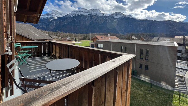 Een houten balkon met een tafel en stoelen, met uitzicht op een bergachtig landschap met besneeuwde toppen en een heldere lucht.
