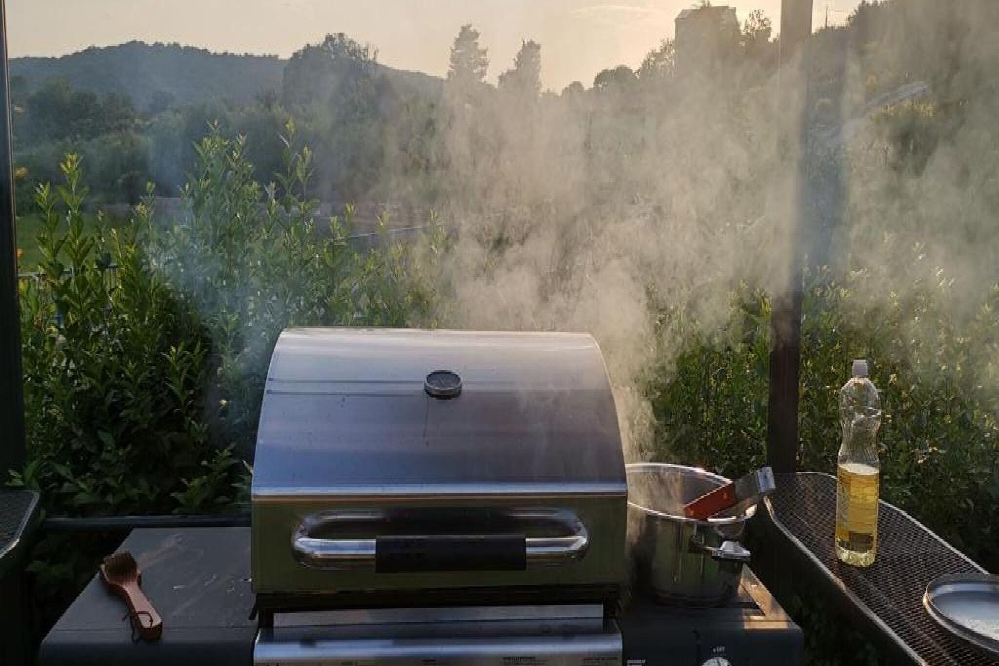 Un barbecue avec une casserole et une bouteille d’huile posés sur une table, le tout sur fond de verdure et de collines.