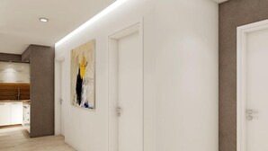 Un pasillo con paredes blancas, suelo de madera y un cuadro. Se observa una puerta a la derecha y una zona de cocina a la izquierda.