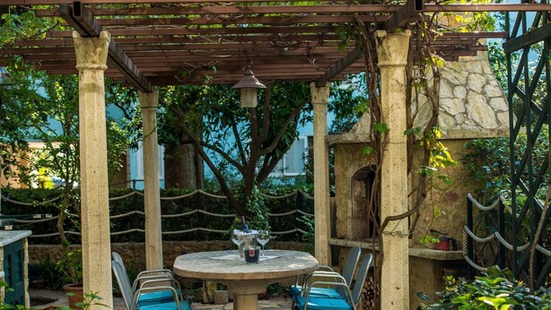 Un patio de piedra con una mesa redonda y cuatro sillas, rodeado de follaje y una estructura de pérgola.