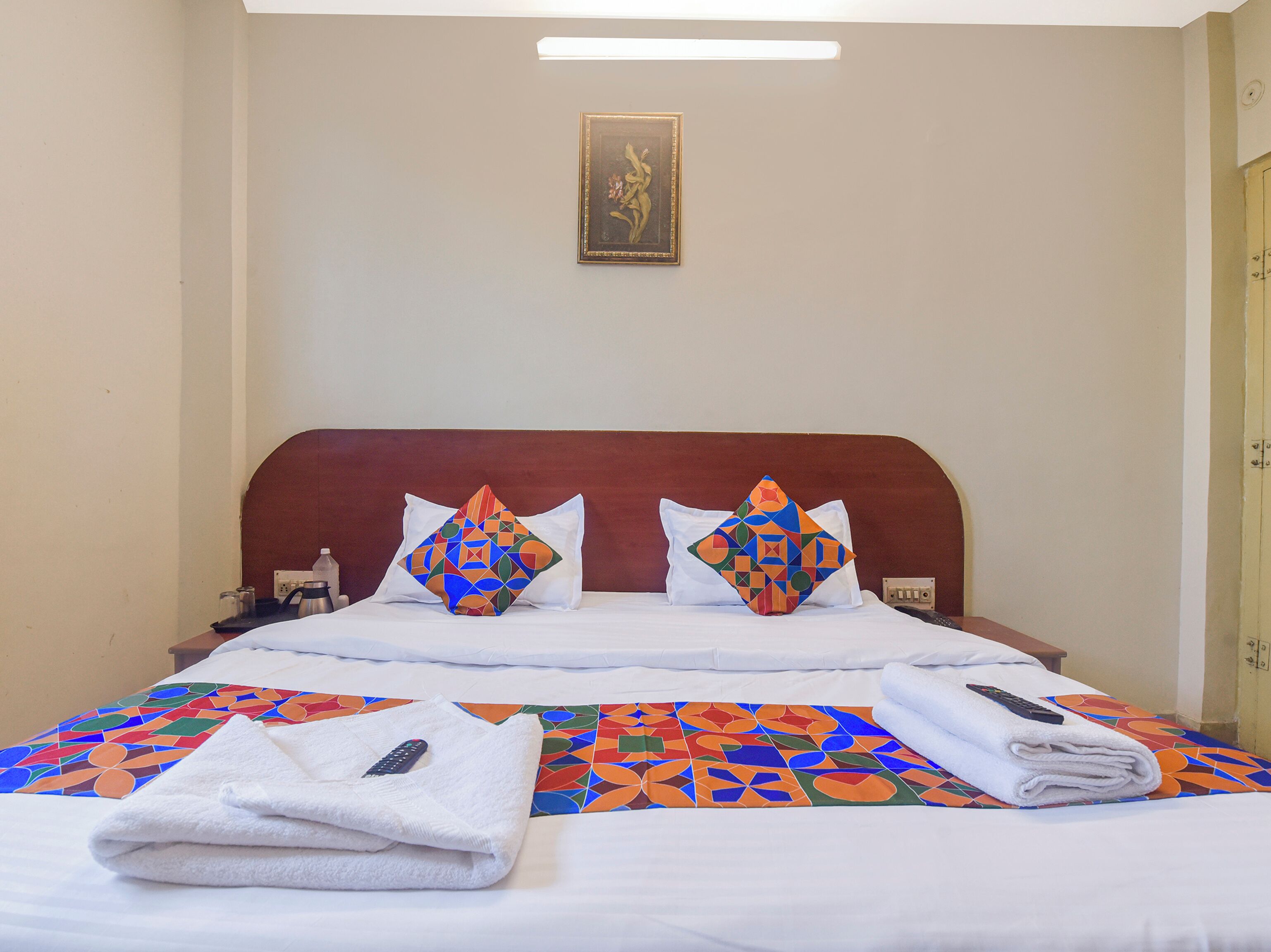 A hotel room with a bed, two pillows, a blanket, a remote control, and a painting on the wall.