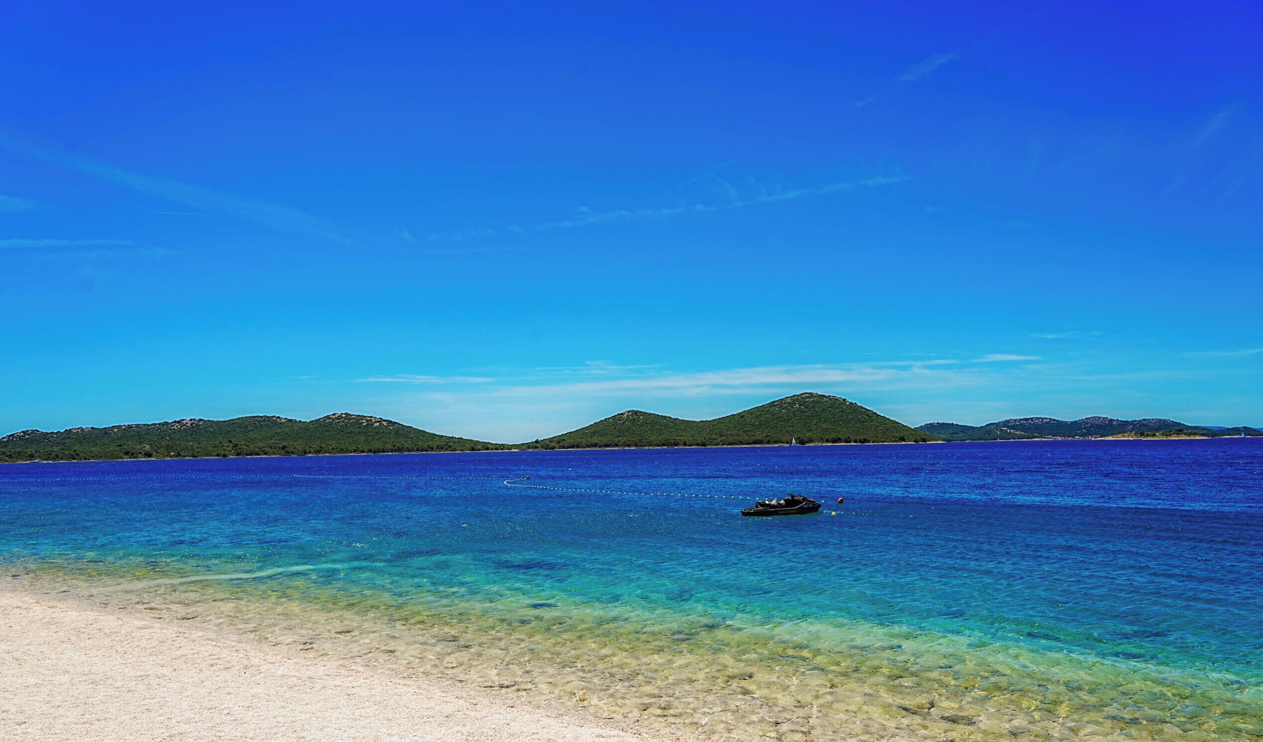 Une plage bordée d’une eau turquoise et limpide, un bateau et, au loin, des collines verdoyantes.