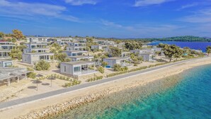 A coastal area with modern, compact housing units arranged along a beach, featuring clear blue waters and a hilly backdrop.