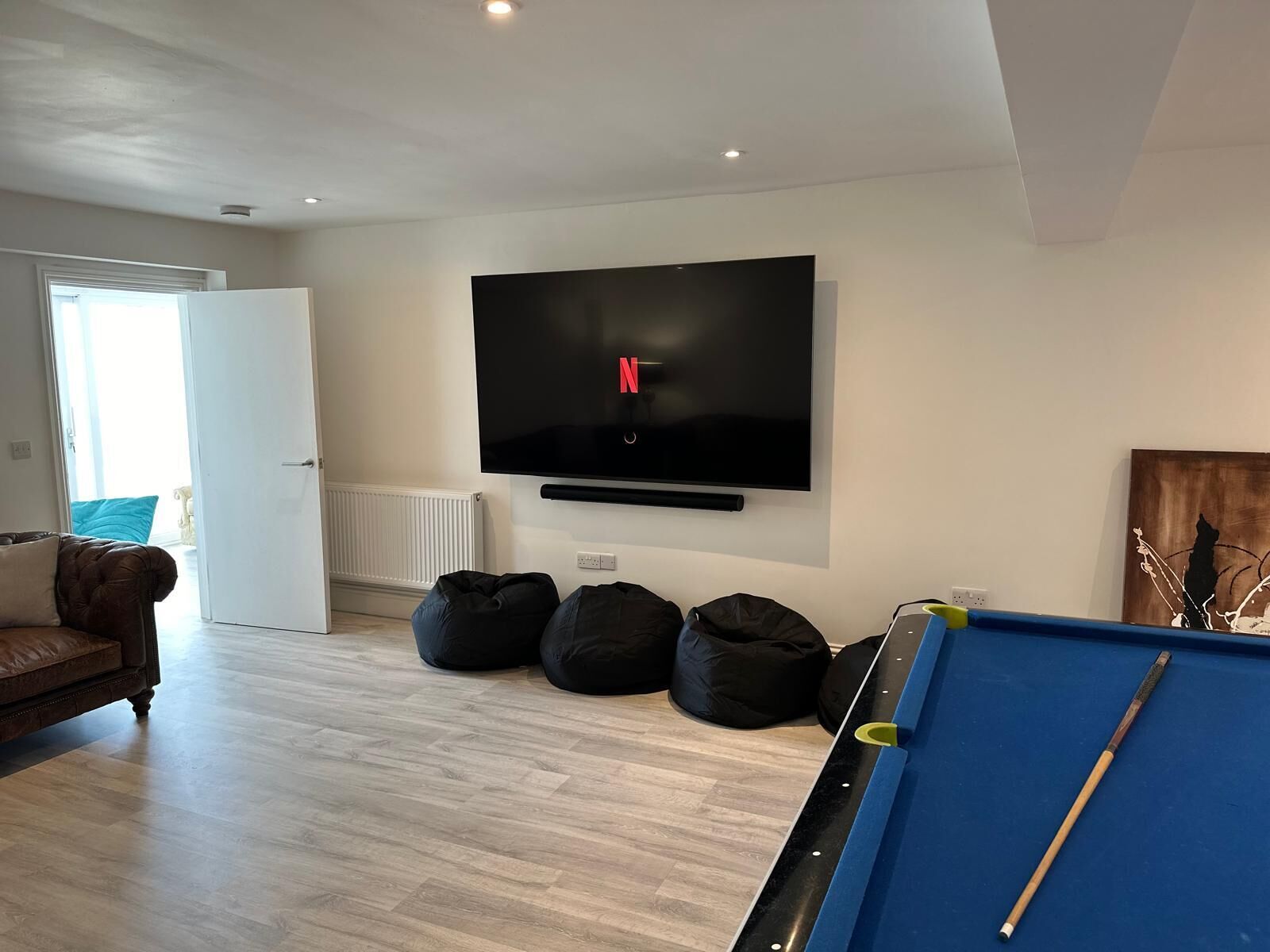 A living room with a pool table, a brown leather sofa, and a flat-screen TV mounted on the wall.