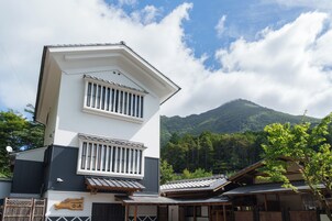 Ein traditionelles japanisches Gebäude mit geneigtem Dach und Holzbalken, umgeben von üppiger Vegetation und einem Berg im Hintergrund.