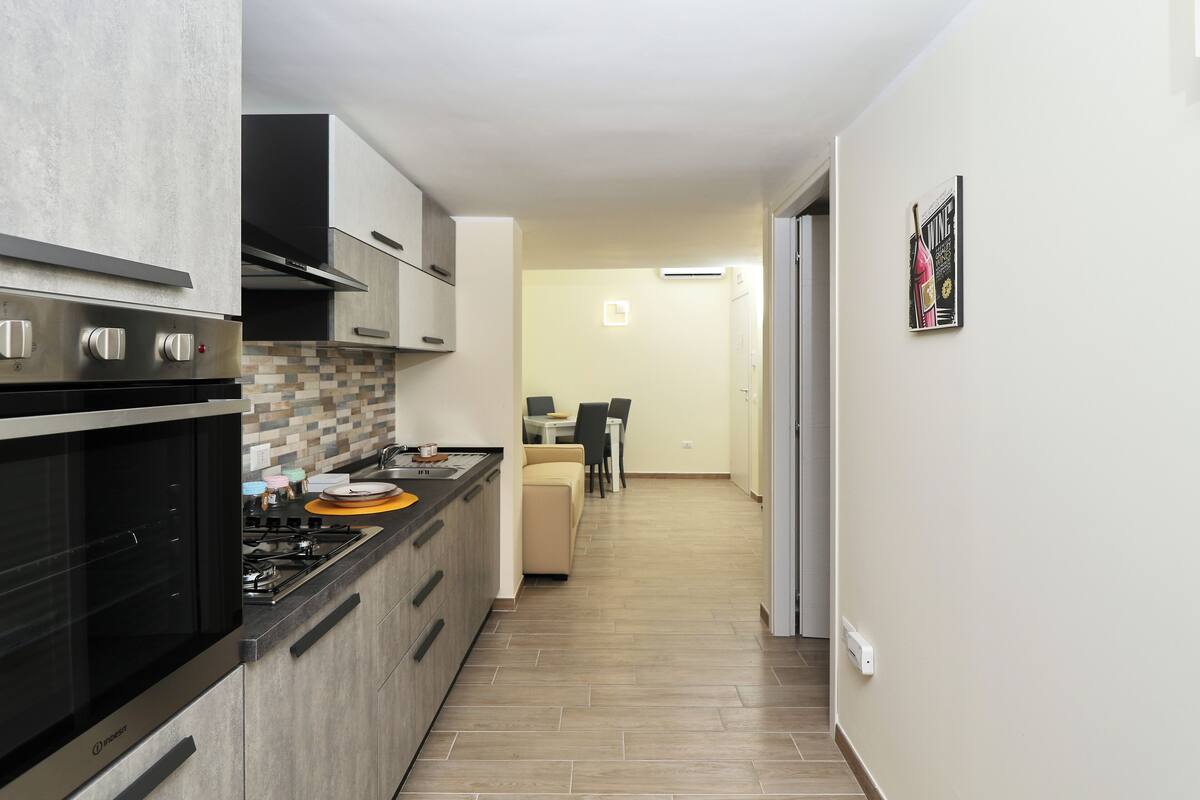 A modern kitchen with stainless steel appliances, a dining area with a sofa, and a hallway leading to a door.