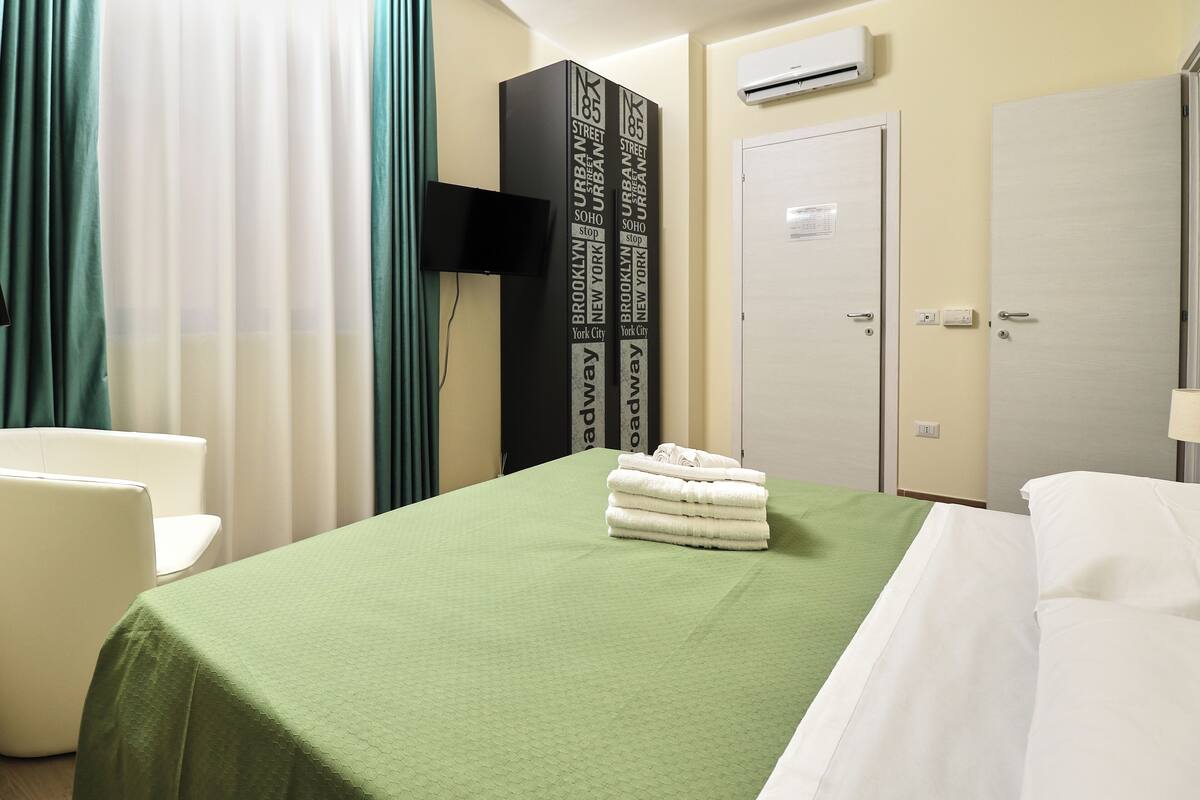 A hotel room with a green bedspread, white pillows, a stack of white towels, a flat-screen TV, and a door with a sign.