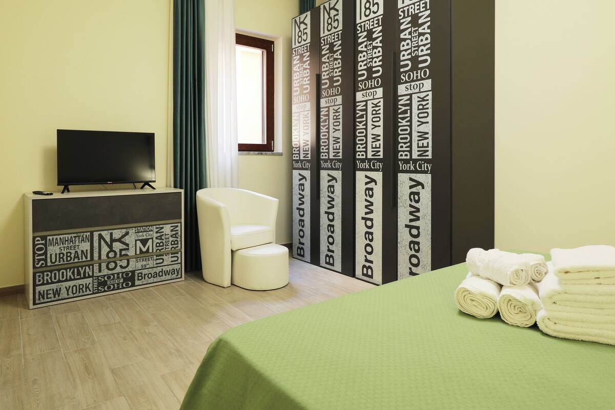 A hotel room with a bed, a TV on a stand, a chair, and a wall with printed text.