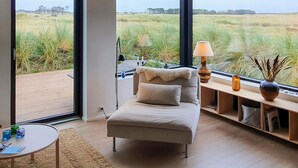 Ein modernes Zimmer mit einem großen Fenster und Blick auf ein grünes Feld. Es gibt ein Sofa mit einem Fellüberwurf, einen kleinen Tisch mit Gläsern sowie eine Lampe auf einem Beistelltisch.
