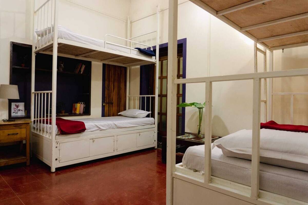 A dormitory room with bunk beds, a small desk with a lamp, and a window with a view of the outside.