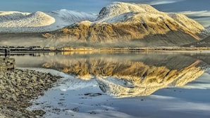 Ein ruhiger See mit schneebedeckten Bergen im Hintergrund und einer Steinstruktur links.