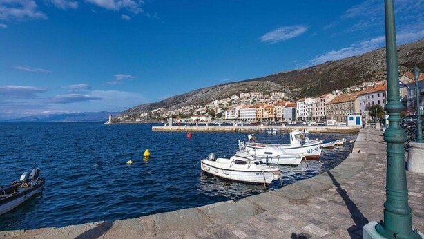 A coastal town with boats docked along a stone quay, buildings in the background, and a clear blue sky.