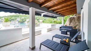 Un espace extérieur aménagé, abrité et doté de mobilier moderne, offrant une vue sur une piscine et sur la verdure, ainsi qu’un mur en pierre.
