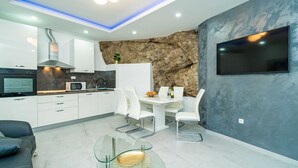 Une cuisine moderne avec des armoires blanches, un four encastré et un four à micro-ondes. On y trouve un coin repas avec une table en verre et des chaises blanches. Un téléviseur à écran plat est fixé au mur.
