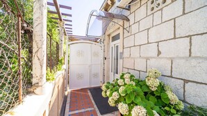 A narrow pathway leading to a white door with decorative panels, flanked by a brick wall and a potted hydrangea plant.