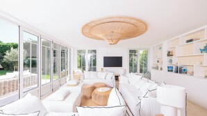 Una sala de estar luminosa y moderna con ventanas amplias, un sofá grande, mesas auxiliares de mimbre y un televisor de pantalla plana montado en la pared.