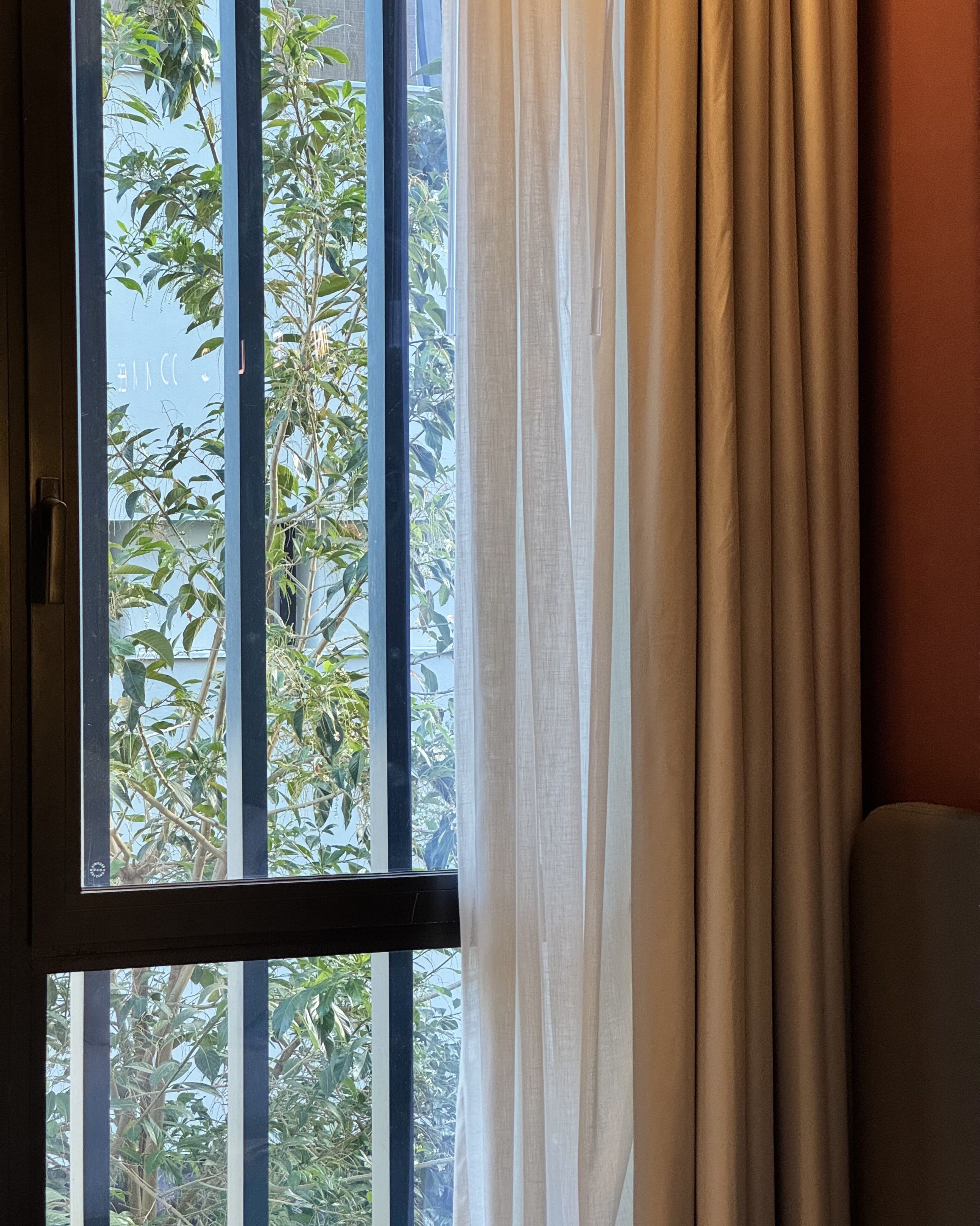 Janela com venezianas verticais, vista para o verde lá fora e cortina parcialmente aberta.