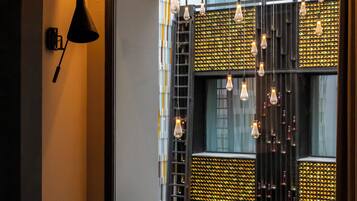 A modern hotel room with a glass table, two chairs, a laptop, a black lamp, and a view of a cityscape through large windows.