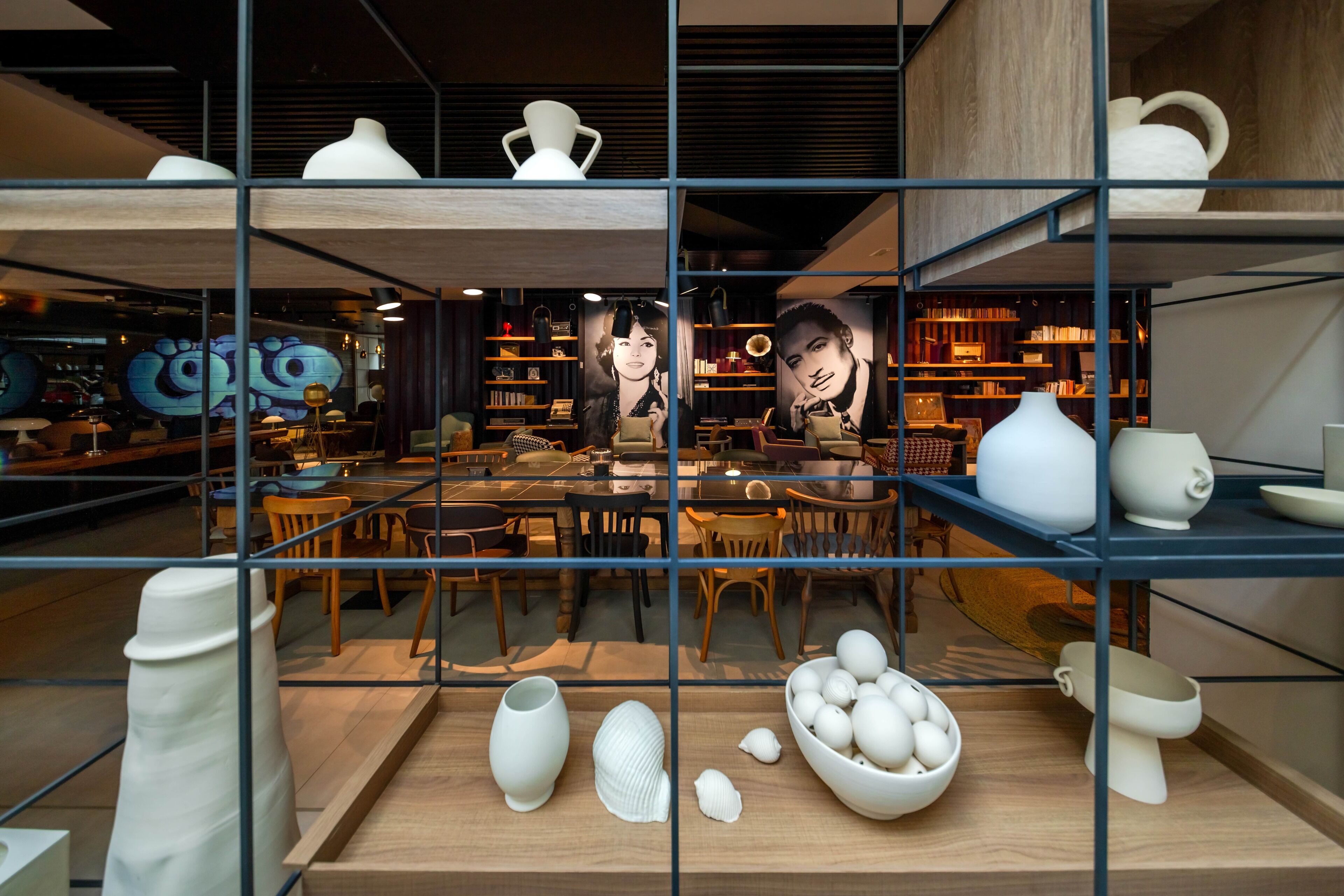 A modern interior with a central table surrounded by chairs, shelves with decorative items, and a glass partition with a view of the dining area.