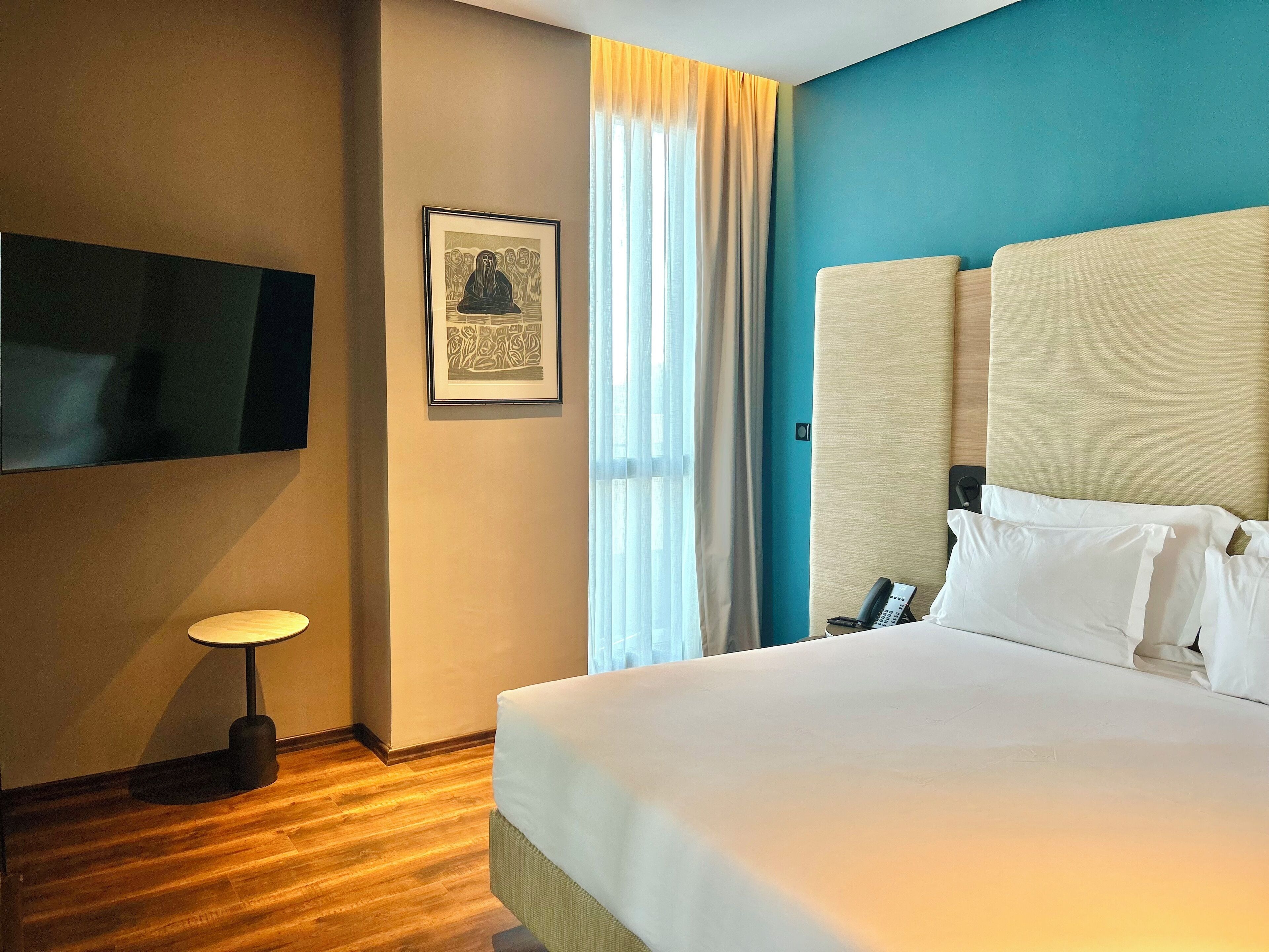 A hotel room with a large bed, a flat-screen TV mounted on the wall, a framed artwork above the bed, a bedside telephone, and a window with curtains.