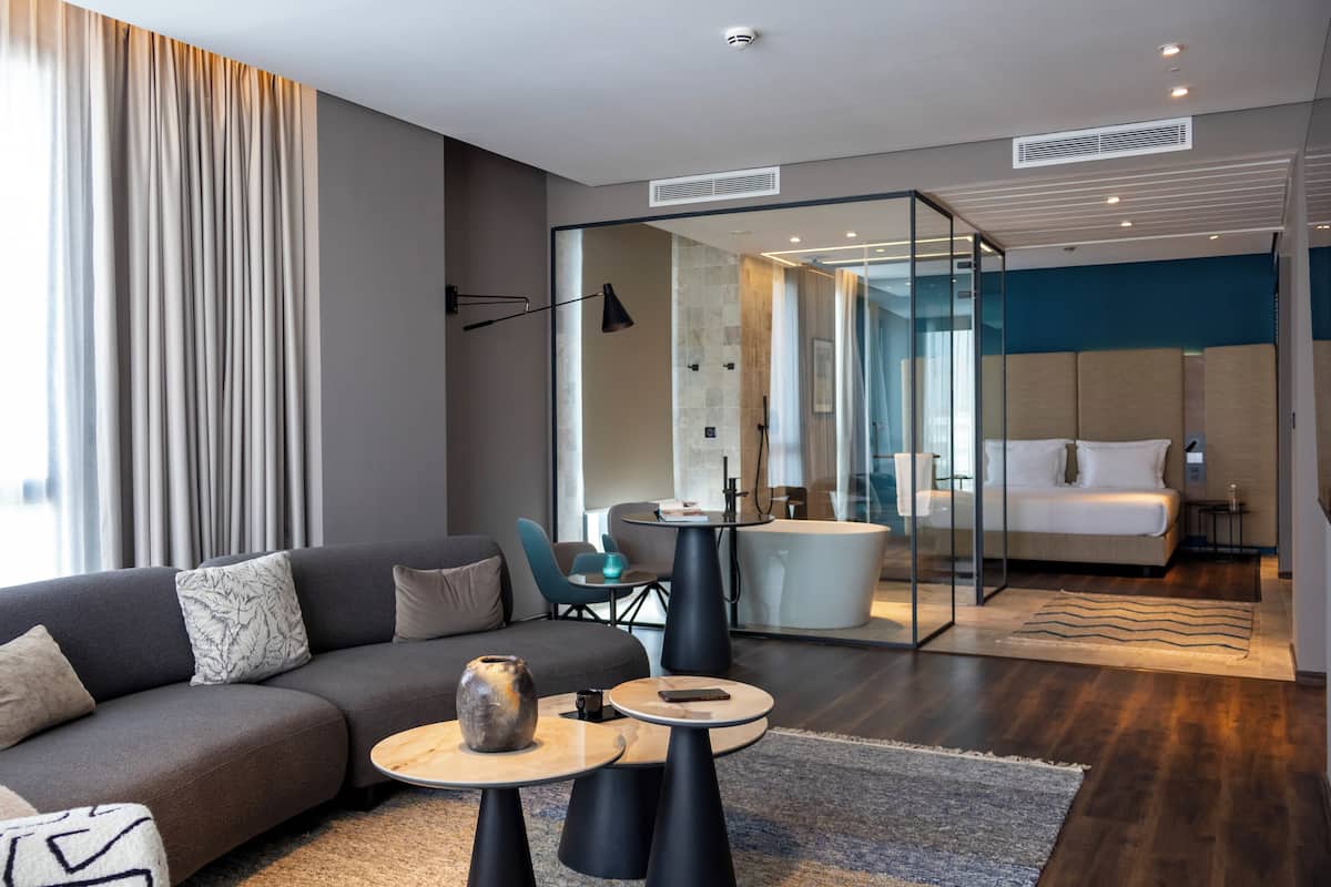 Ein modernes Hotelzimmer mit Sofa, Couchtisch und einem durch eine Glasscheibe abgetrennten Badezimmer.