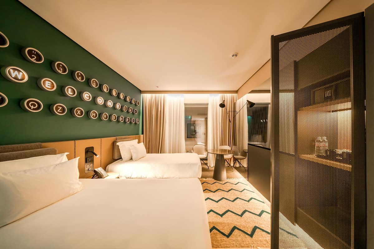 Ein Hotelzimmer mit zwei Betten, einer Wand mit beleuchteten Buchstaben, einem kleinen Tisch mit Stühlen und Blick durch eine Tür.