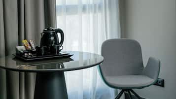 Quarto de hotel moderno com uma mesa de vidro redonda, uma cadeira cinza, um abajur preto e uma janela com cortinas translúcidas.