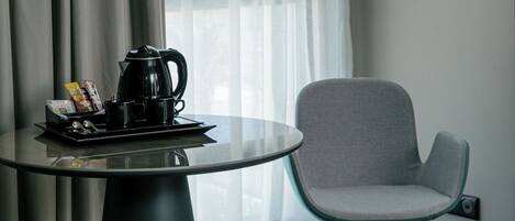 A modern hotel room with a round glass table, a grey chair, a black floor lamp, and a window with sheer curtains.