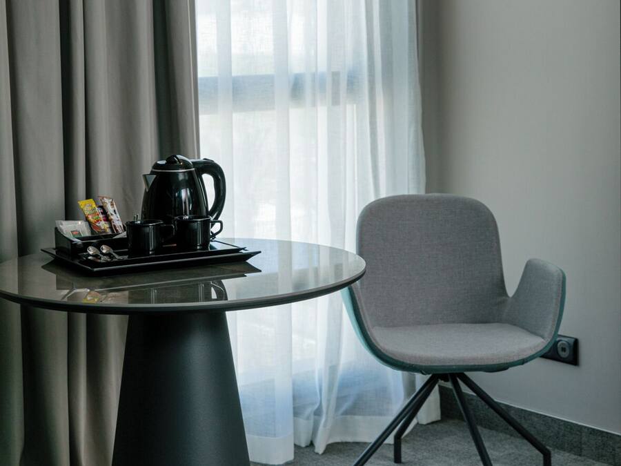 Quarto de hotel moderno com uma mesa de vidro redonda, uma cadeira cinza, um abajur preto e uma janela com cortinas translúcidas.