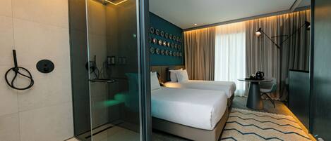 A modern hotel room with a glass shower enclosure, a bed with two pillows, a small round table with a coffee pot, and a wall-mounted control panel.