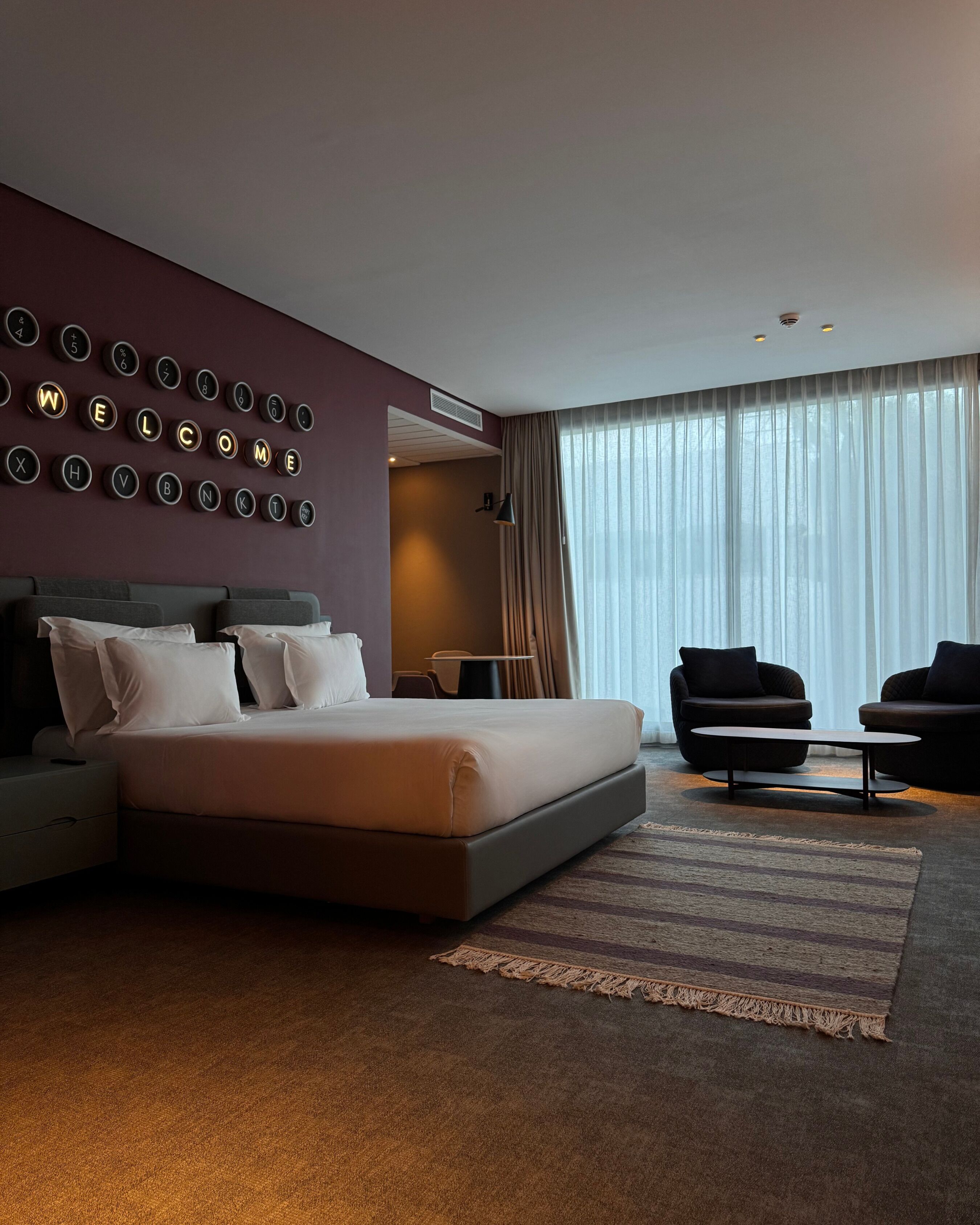 Une chambre d’hôtel moderne avec un grand lit, deux fauteuils, une petite table et un mur orné de lettres lumineuses qui forment le mot « BIENVENUE ».