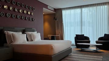 Une chambre d’hôtel moderne avec un grand lit, deux fauteuils, une petite table et un mur orné de lettres lumineuses qui forment le mot « BIENVENUE ».