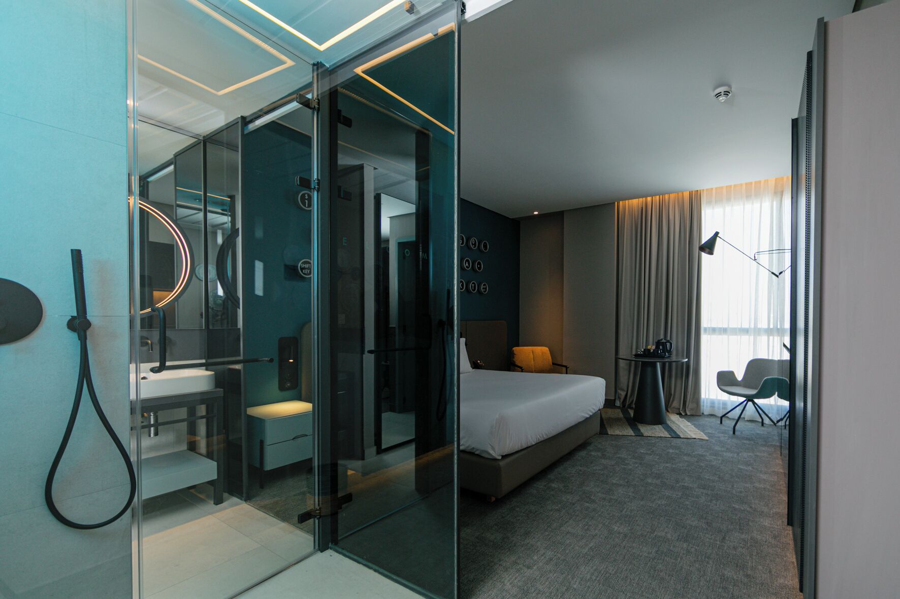 A modern hotel room with a glass partition, a bed, a desk, and a chair.