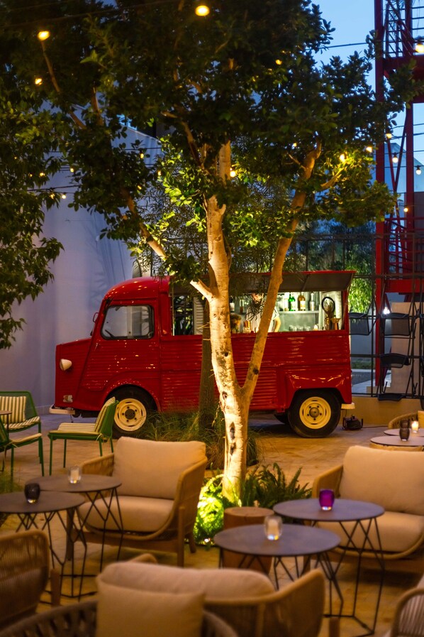 A red vintage car converted into a food truck, surrounded by outdoor seating and a tree. - The NINE Lifestyle Experience (La Marsa)