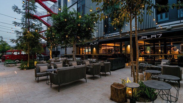 An outdoor seating area with wicker furniture and string lights, a red fire escape, and a building with a 'LA PLA7' sign. - The NINE Lifestyle Experience (La Marsa)