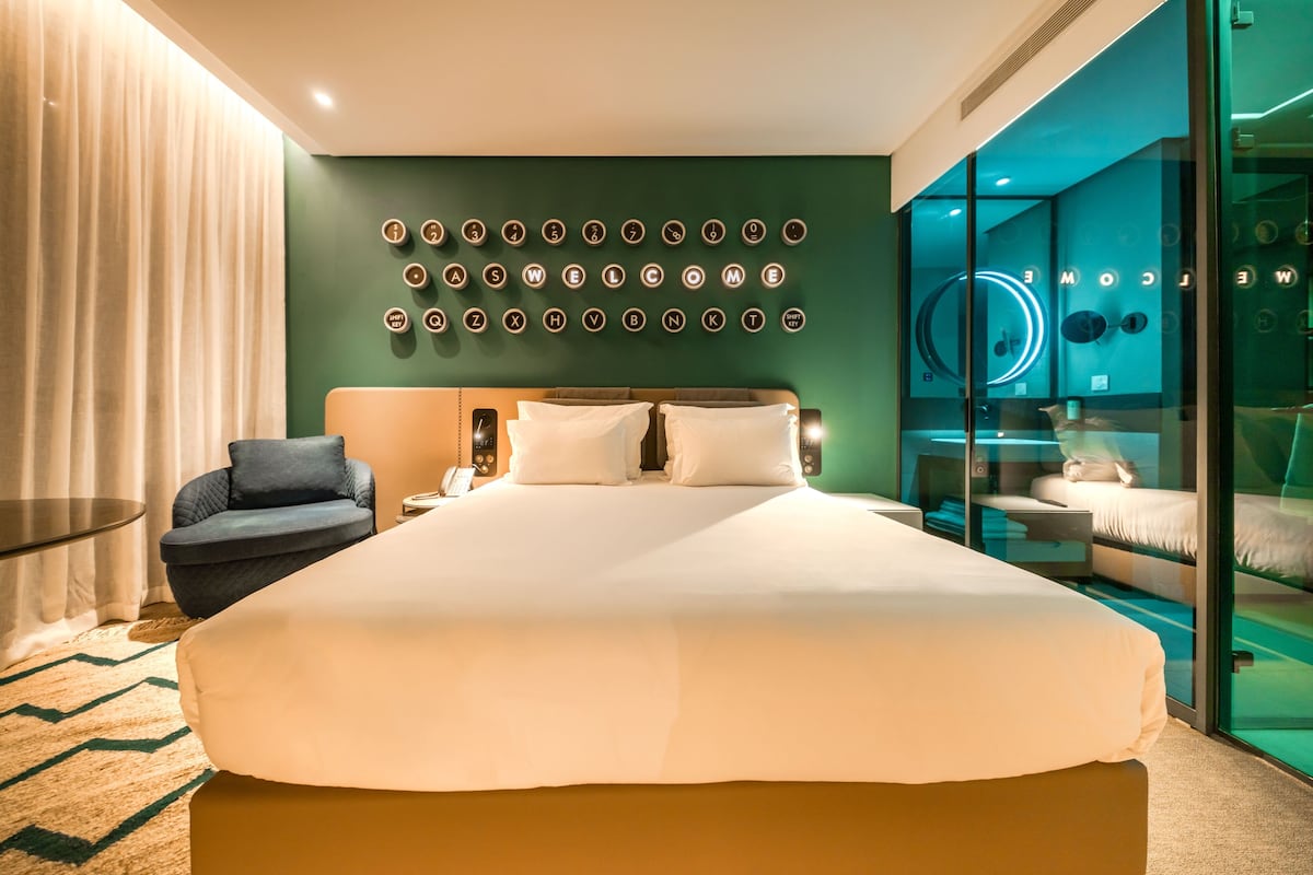 Ein modernes Hotelzimmer mit einem großen Bett, einer Glastrennwand, einer Sitzecke und einer Wand mit beleuchteten Buchstaben.
