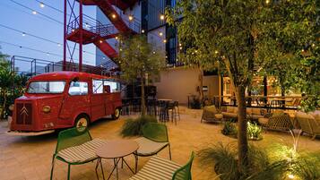 Una furgoneta roja antigua estacionada en un patio con mesas al aire libre, rodeada de árboles y luces colgantes.