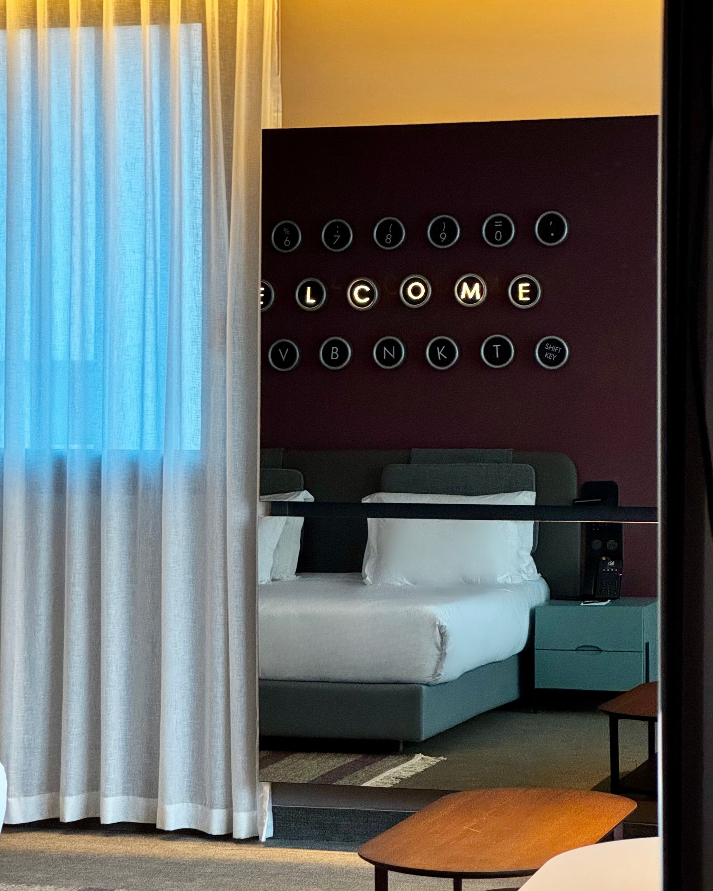 A hotel room with a bed, bedside tables, a wall-mounted control panel, and a window with curtains.