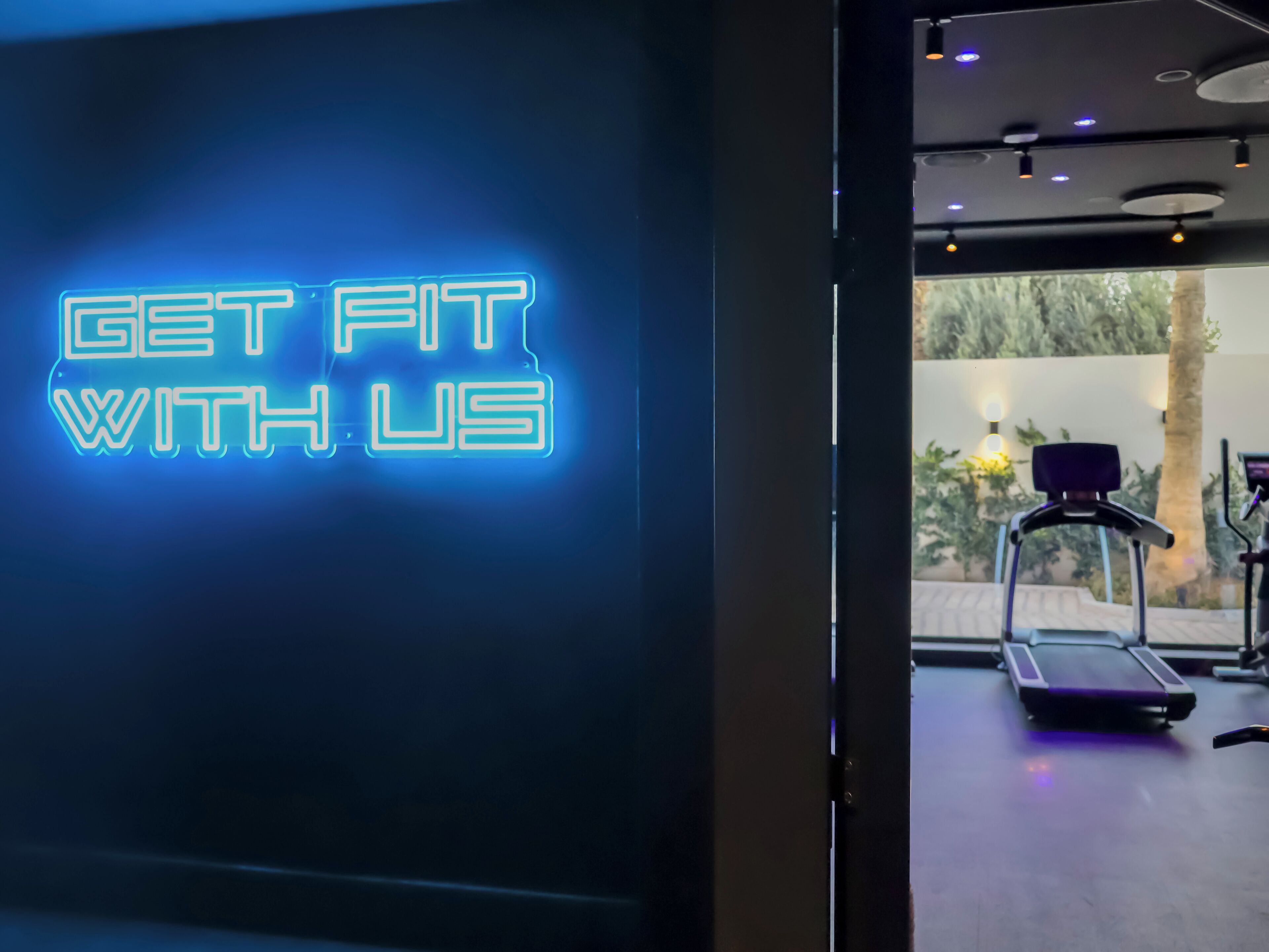 L’intérieur d’une salle de sport, avec un panneau néon sur lequel on peut lire « ENTRAÎNEZ-VOUS AVEC NOUS » et divers équipements d’exercice.
