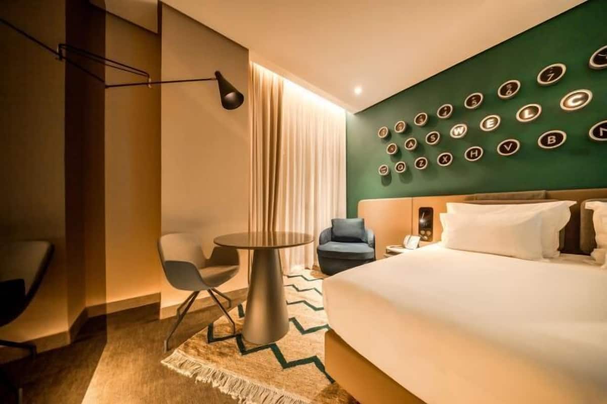 Ein Hotelzimmer mit Bett, rundem Tisch, Stuhl, Schreibtisch, Lampe, Wanduhr und Vorhang.
