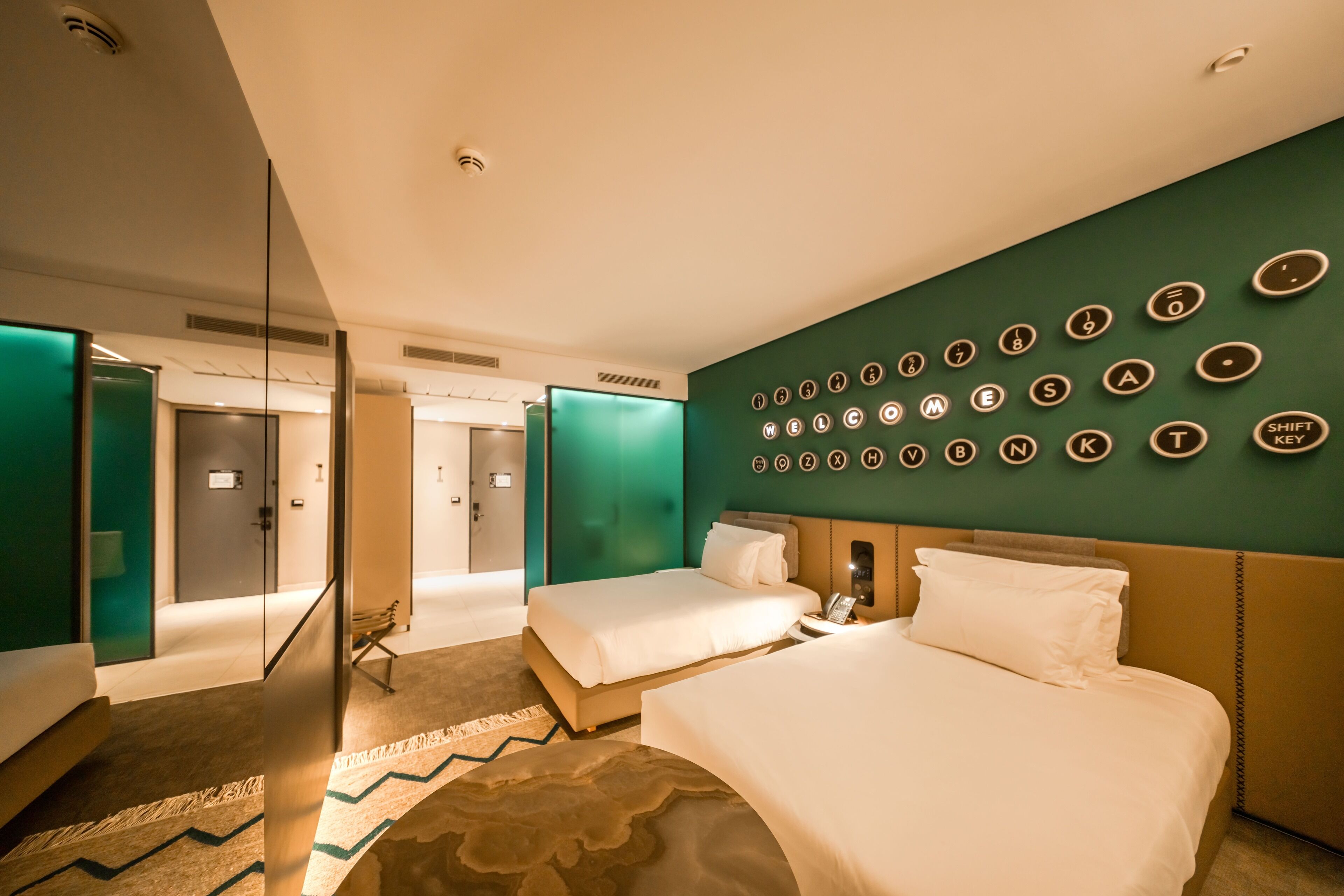 Habitación de hotel con dos camas, una pared de acento verde, un teclado montado en la pared y un panel de vidrio.
