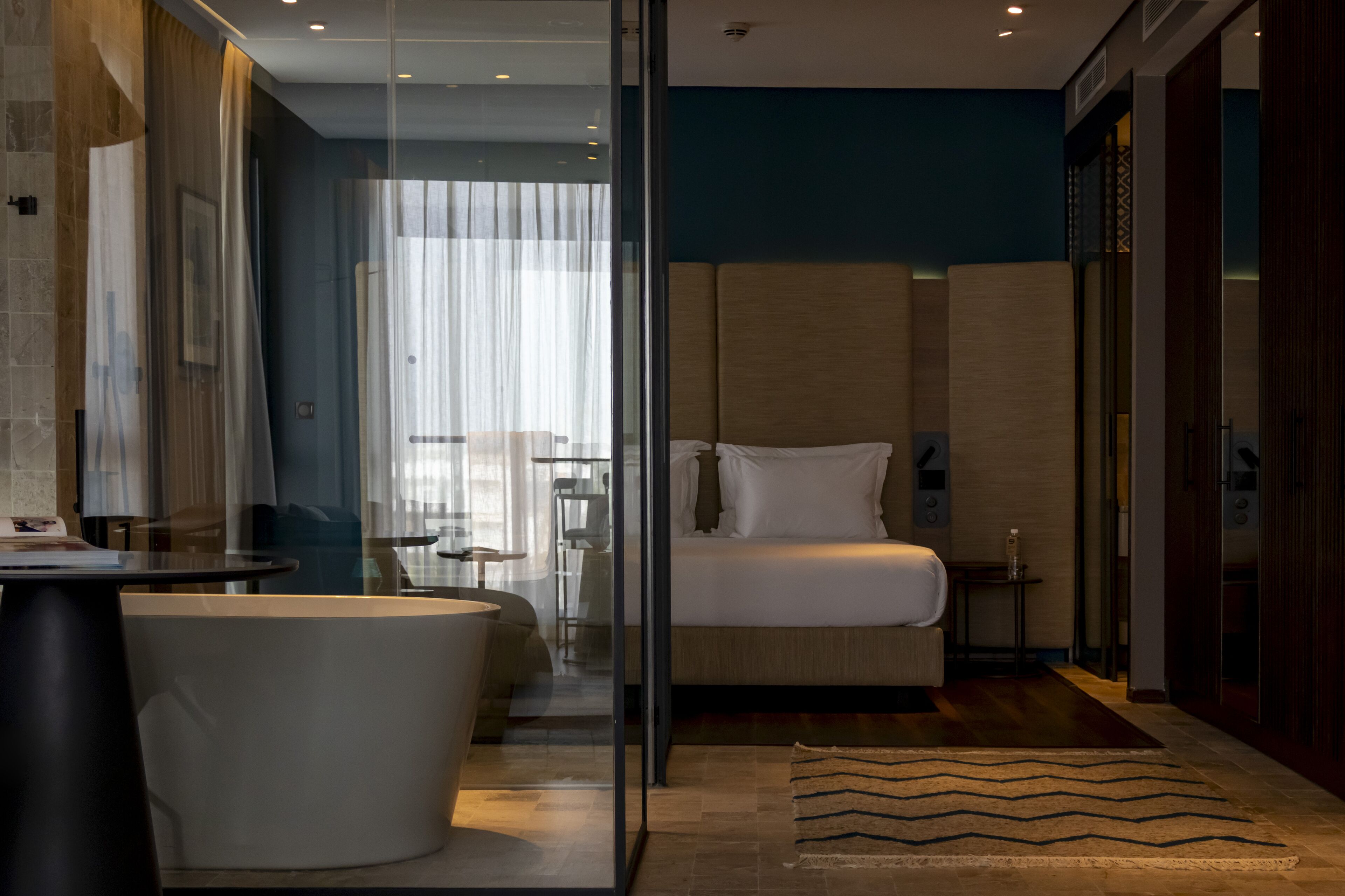Una moderna camera d'albergo con cabina doccia in vetro, un letto grande, un'area salotto con una sedia e un tavolo, e vista sulla città dalla finestra.