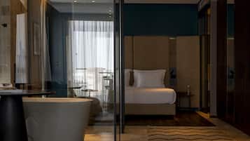 Una habitación de hotel moderna con cabina de ducha de vidrio, una cama grande, un área de estar con una silla y una mesa, y vista a la ciudad por la ventana.