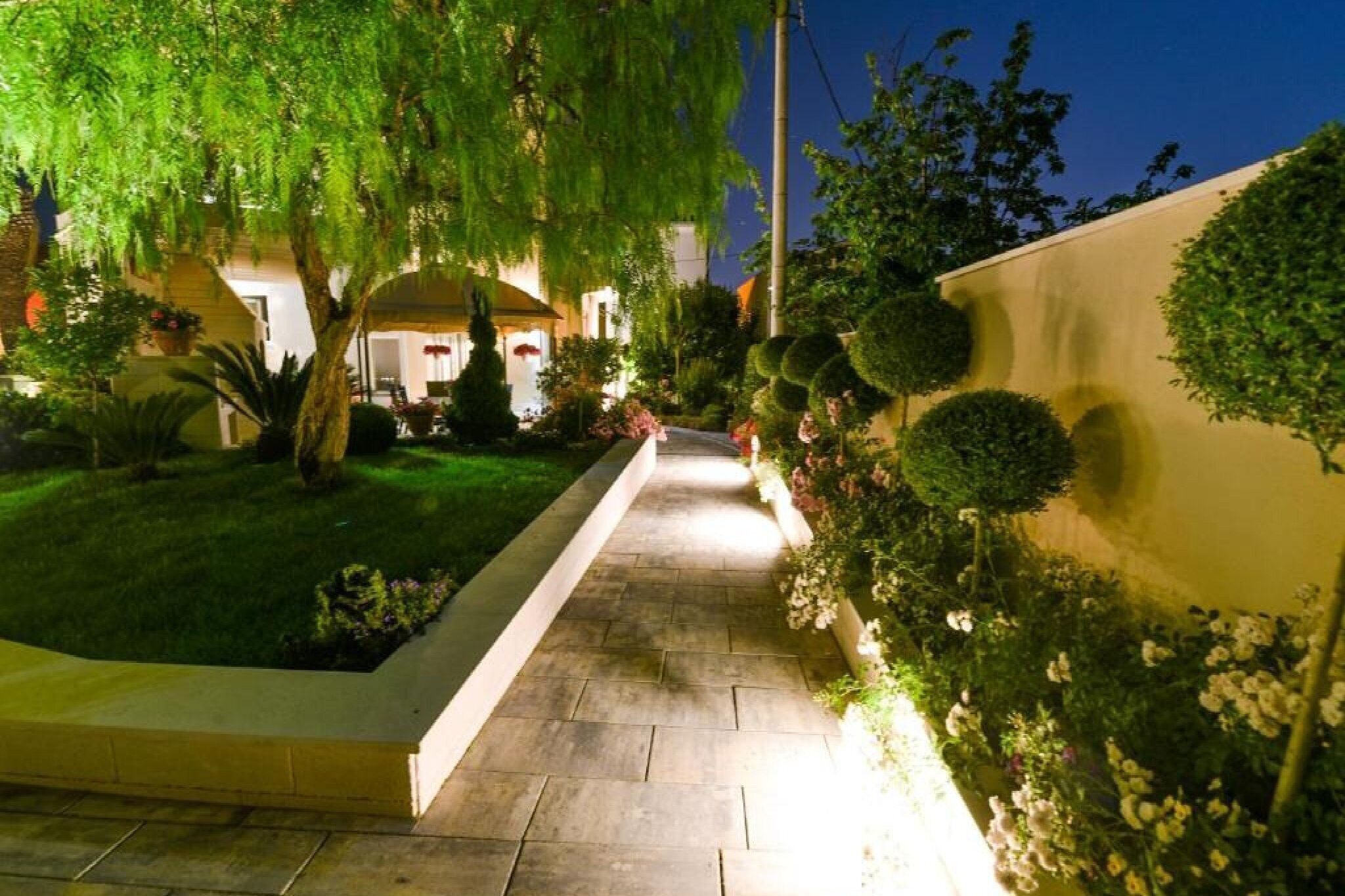 A well-lit pathway leading to a building with a covered entrance, surrounded by neatly trimmed hedges and flowering plants.