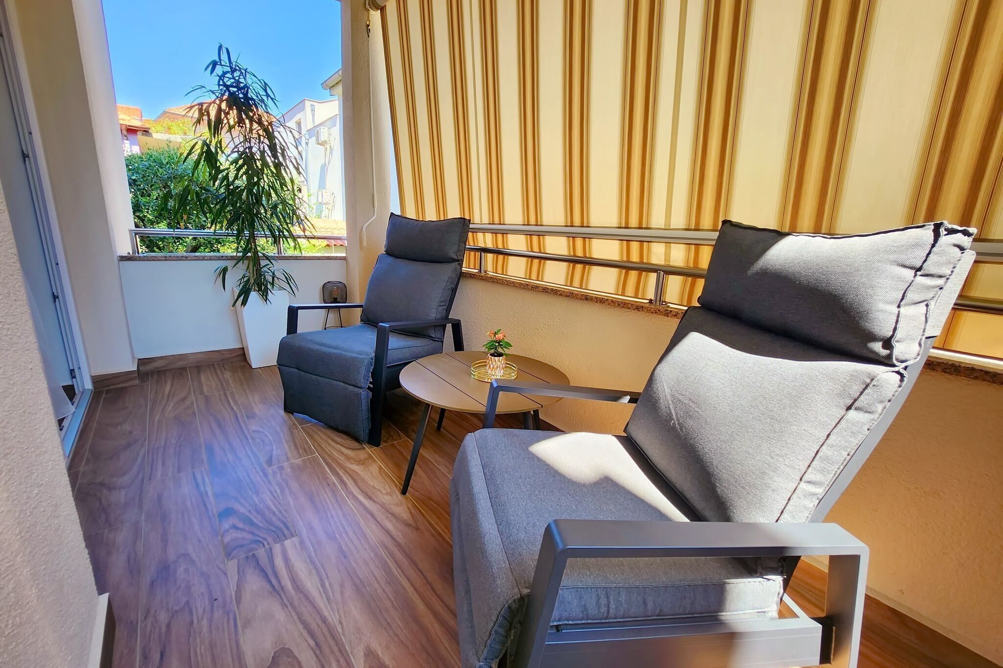 Ein Balkon mit einer Couch und Sesseln, ein kleiner Tisch mit einer Pflanze und Blick auf Grünflächen und Gebäude.