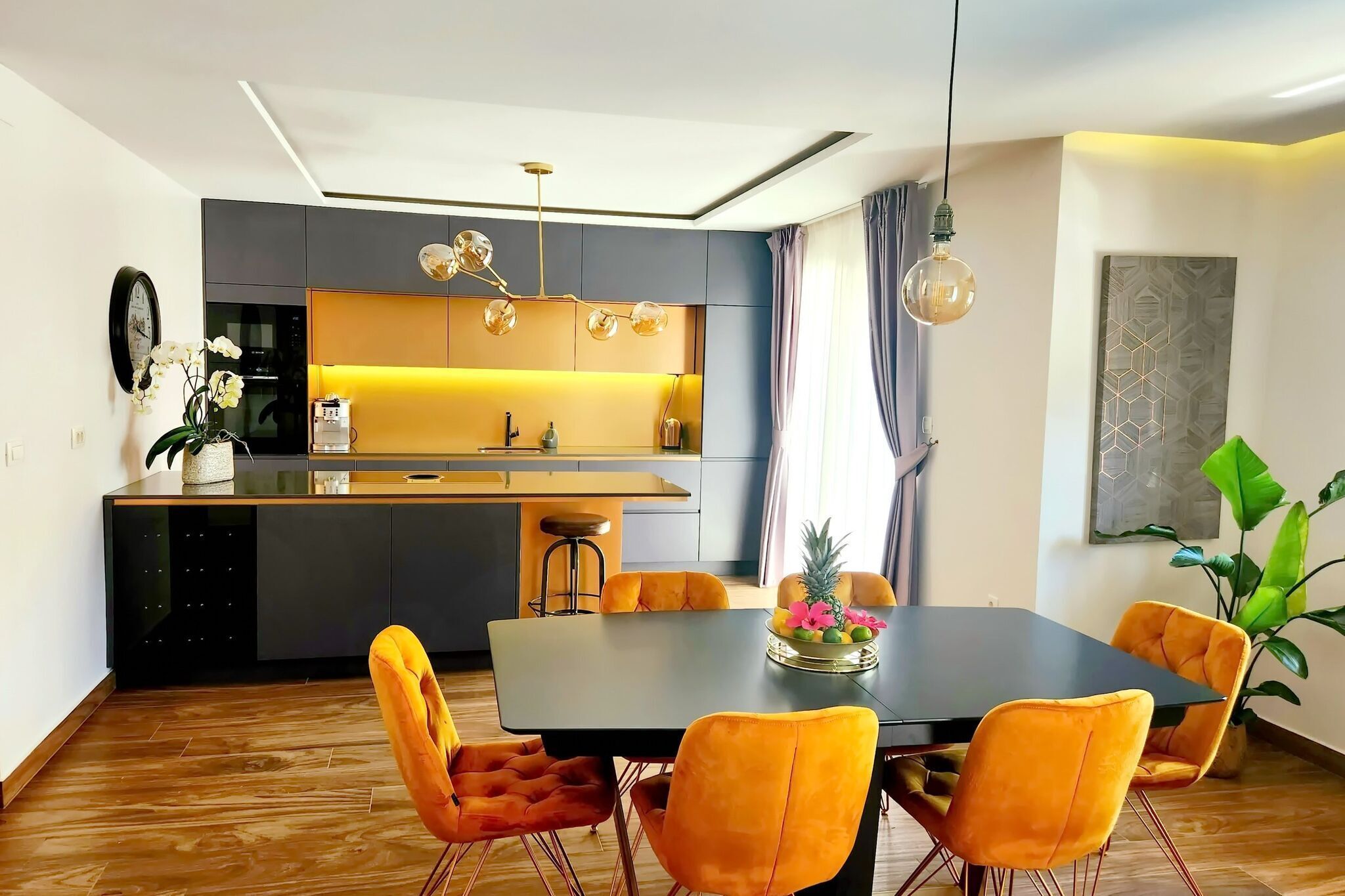 A modern kitchen with a central island, a dining area with a black table and orange chairs, and a large plant in the corner.