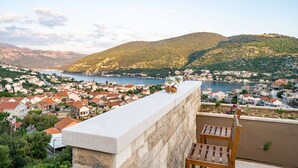 A balcony with a stone balustrade overlooking a coastal town with red-roofed buildings and a view of the sea and mountains.