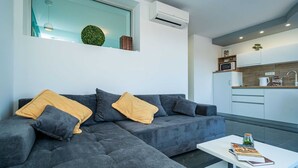 A modern living room with a grey sectional sofa, a white coffee table, and a kitchen area with white cabinets and a microwave.