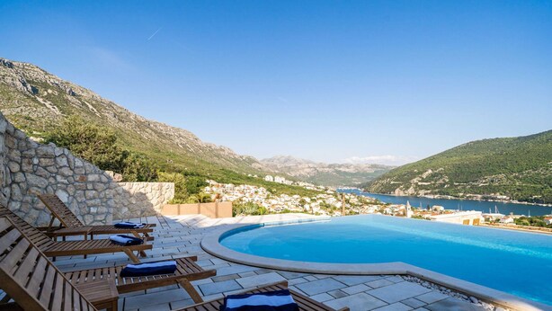 A poolside area with lounge chairs and a stone wall, overlooking a coastal town and mountains.