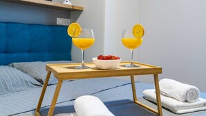 A neatly made bed with a blue headboard, a wooden bedside table with two glasses of orange juice and a bowl of strawberries, and folded white towels.